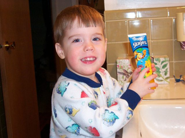 A young boy stands next to a white sink in dinosaur pajamas while holding toddler toothpaste in Penfield, NY