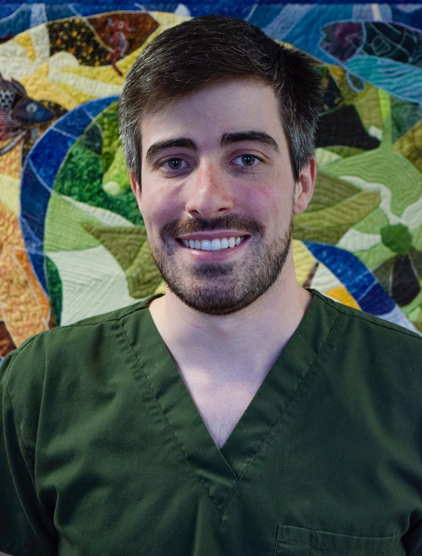 Dr. Benjamin Crilly smiling and wearing green scrubs while standing in front of a mural at Terrance J. O'Keefe, DDS, LLC in Penfield, NY
